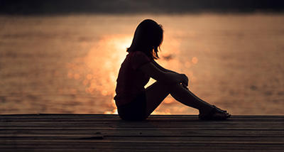 a shadowed woman sitting in a sad posture near water at sunset