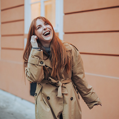 a lady with ginger hair and a brown coat smiling and laughing on the phone