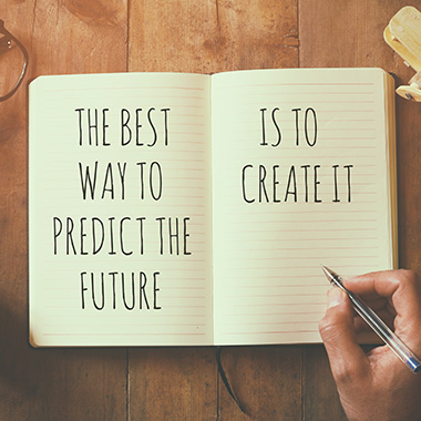 a person writing 'The Best Way to Predict the Future, Is to Create it' in a notebook