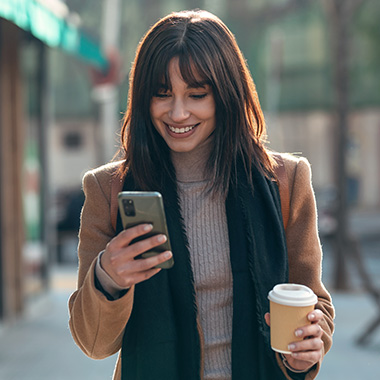 a lady holding her phone with one hand and a hot beverage in the other while smiling and looking at her phone