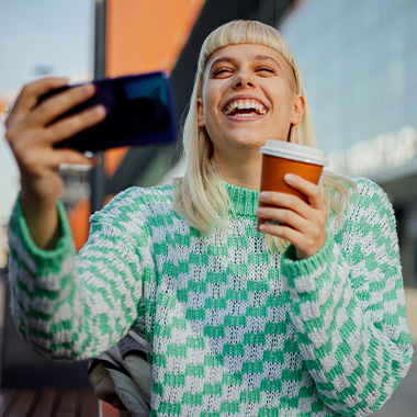 a laughing lady with a green and white jumper holding her phone in one hand and a hot beverage in another